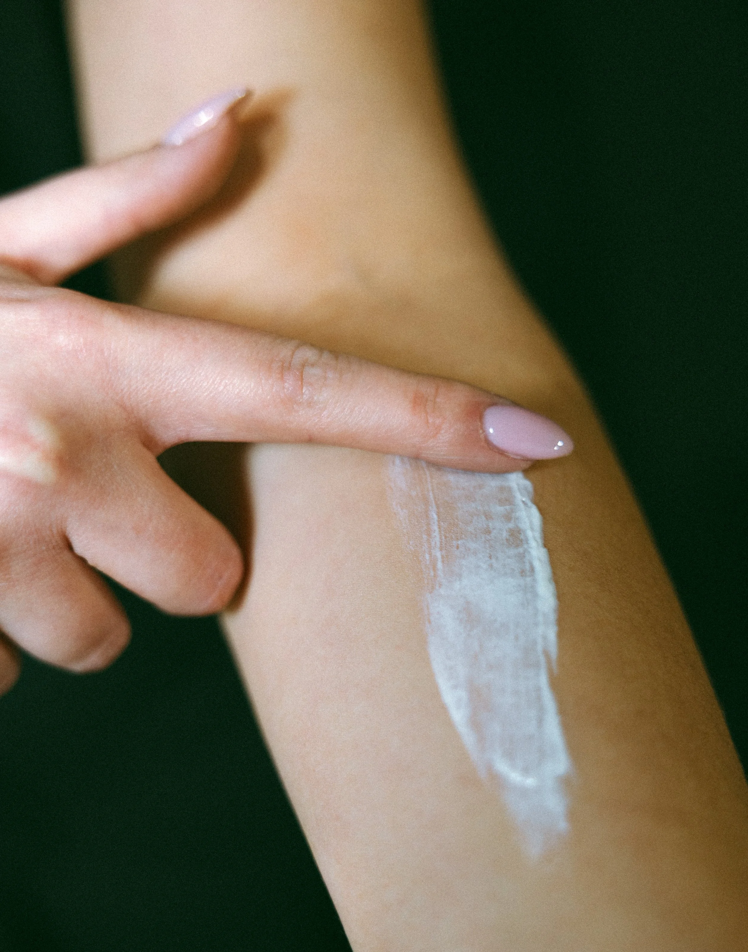 Close-up of a person's hand with manicured nails, applying or touching white cream or lotion on their skin.
