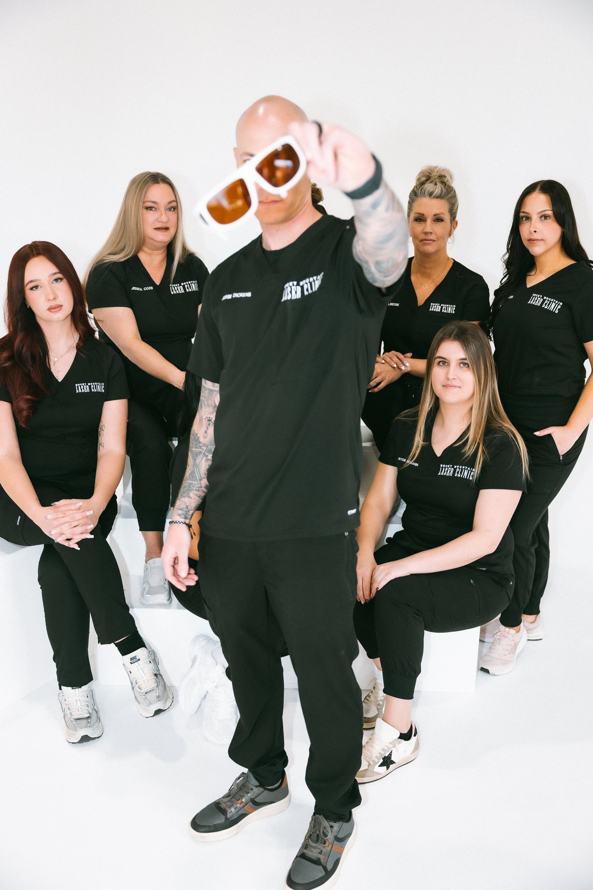 A group of six women and one man posing against a white background, with the man in front holding sunglasses. The women are wearing black shirts with white text, black pants, and sneakers.