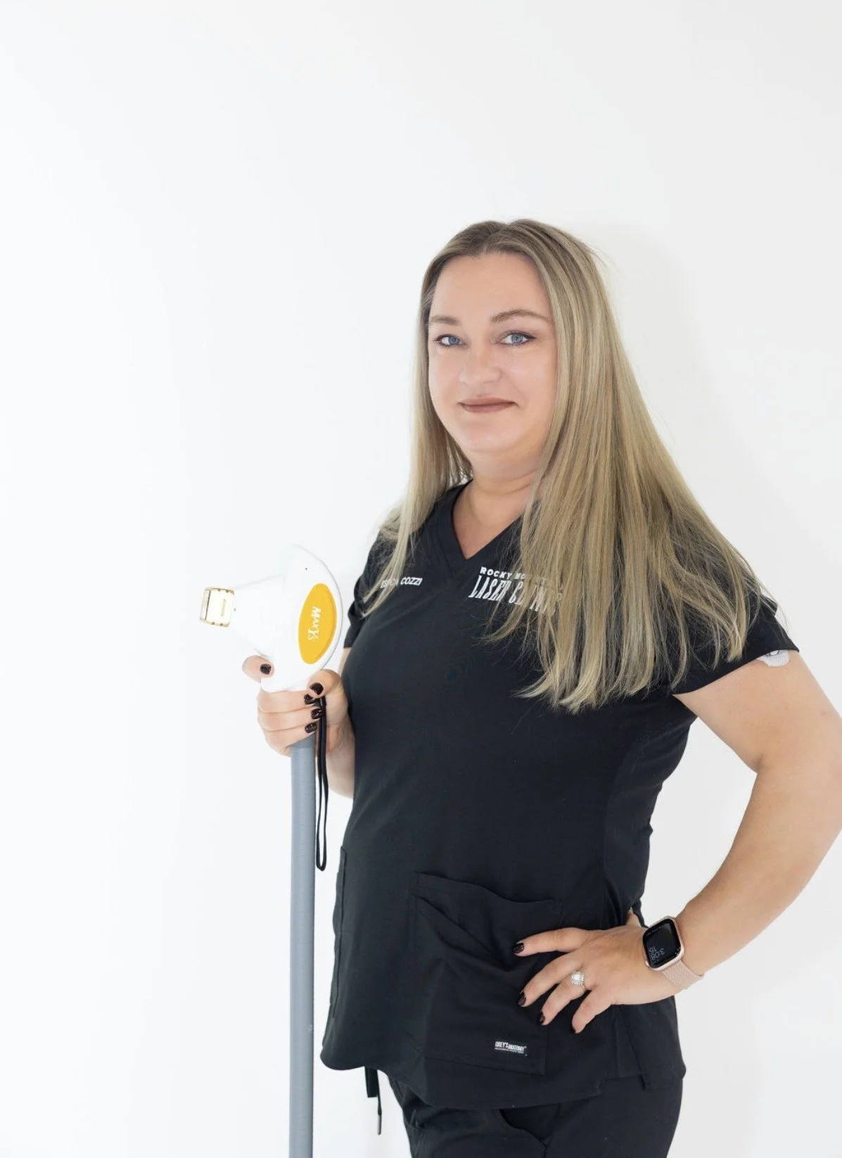 A woman with long blonde hair, wearing a black medical scrub top, holding an ultrasound wand, standing against a plain white background.