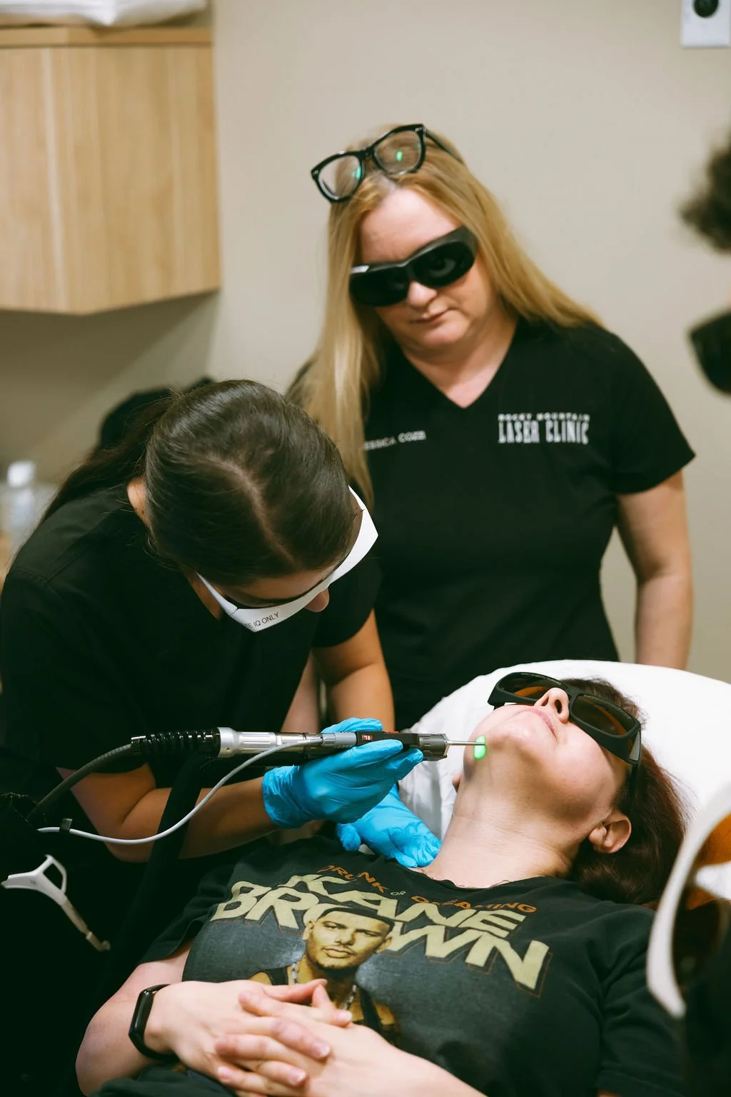 A woman lying on a medical chair is receiving a cosmetic treatment on her chin using a laser device. She is wearing protective glasses. Two professionals are attending to her, one wearing glasses on her head and a mask, and the other in a black T-shirt with a printed logo, both wearing gloves.