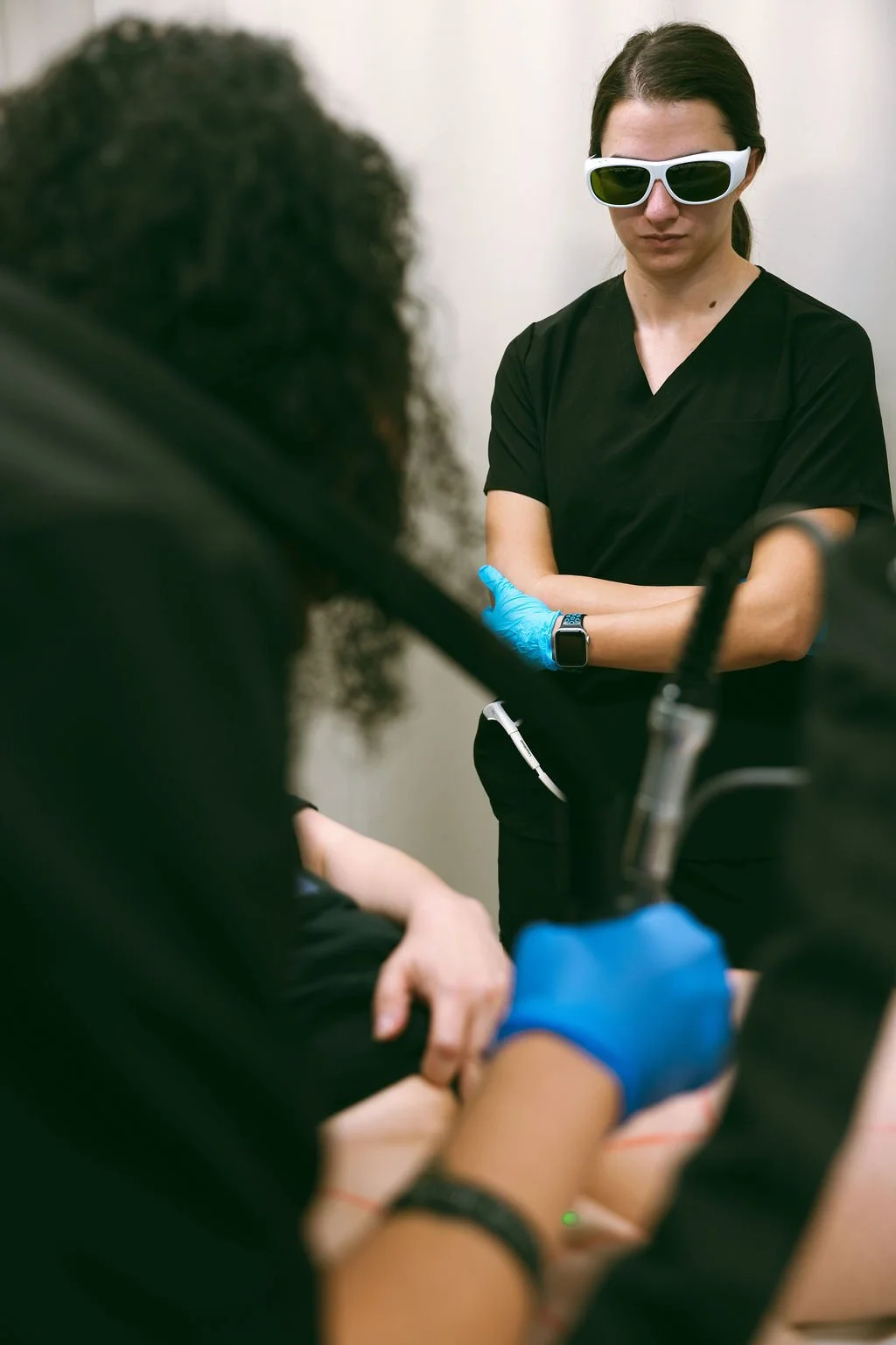 Medical professional wearing sunglasses and a black uniform, standing with arms crossed, observing a patient during a procedure while two other healthcare workers assist.