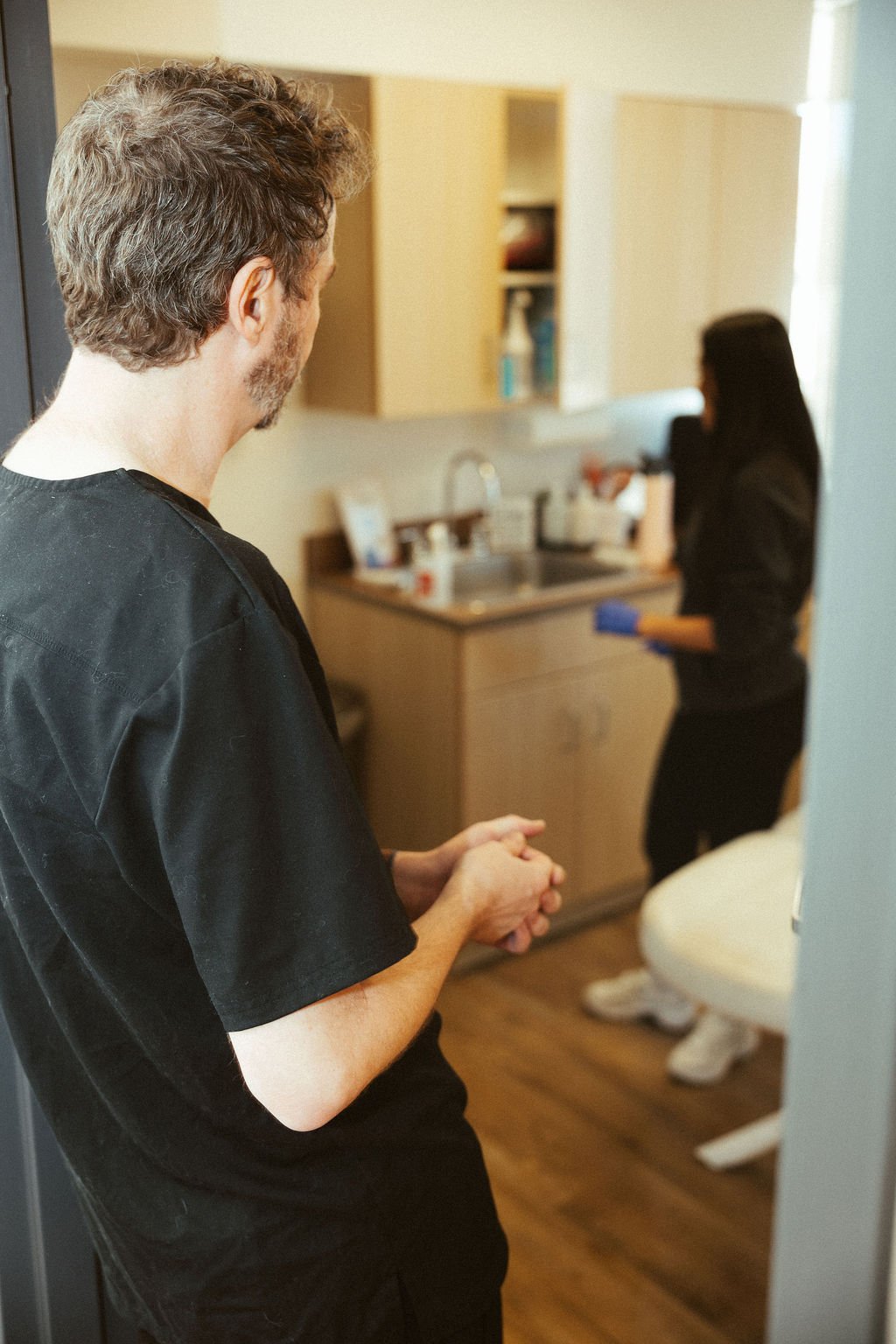 A male healthcare worker with curly hair, wearing black scrubs, standing with clasped hands in a medical setting. In the background, a female healthcare worker with long dark hair, dressed in black, is preparing medical supplies at a countertop in a clinical room.