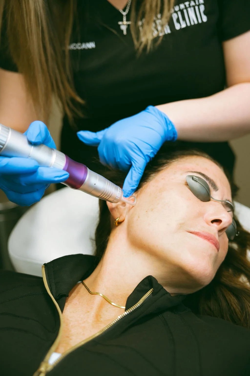 A woman receiving a cosmetic treatment on her face with a handheld device, lying down with eye protection, while a practitioner in blue gloves administers the procedure in a clinical setting.