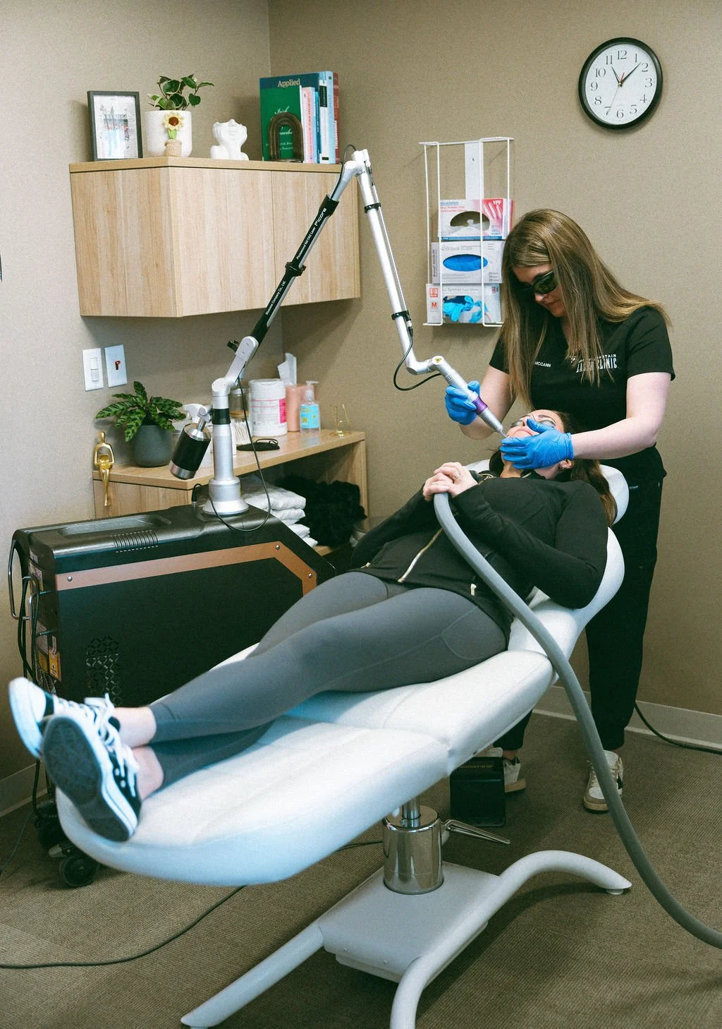 A woman is receiving a dental or medical procedure, lying on a white examination chair, with a healthcare professional wearing black scrubs and blue gloves performing the procedure using a handheld device. The room has beige walls, cabinets, a clock, and various medical supplies.