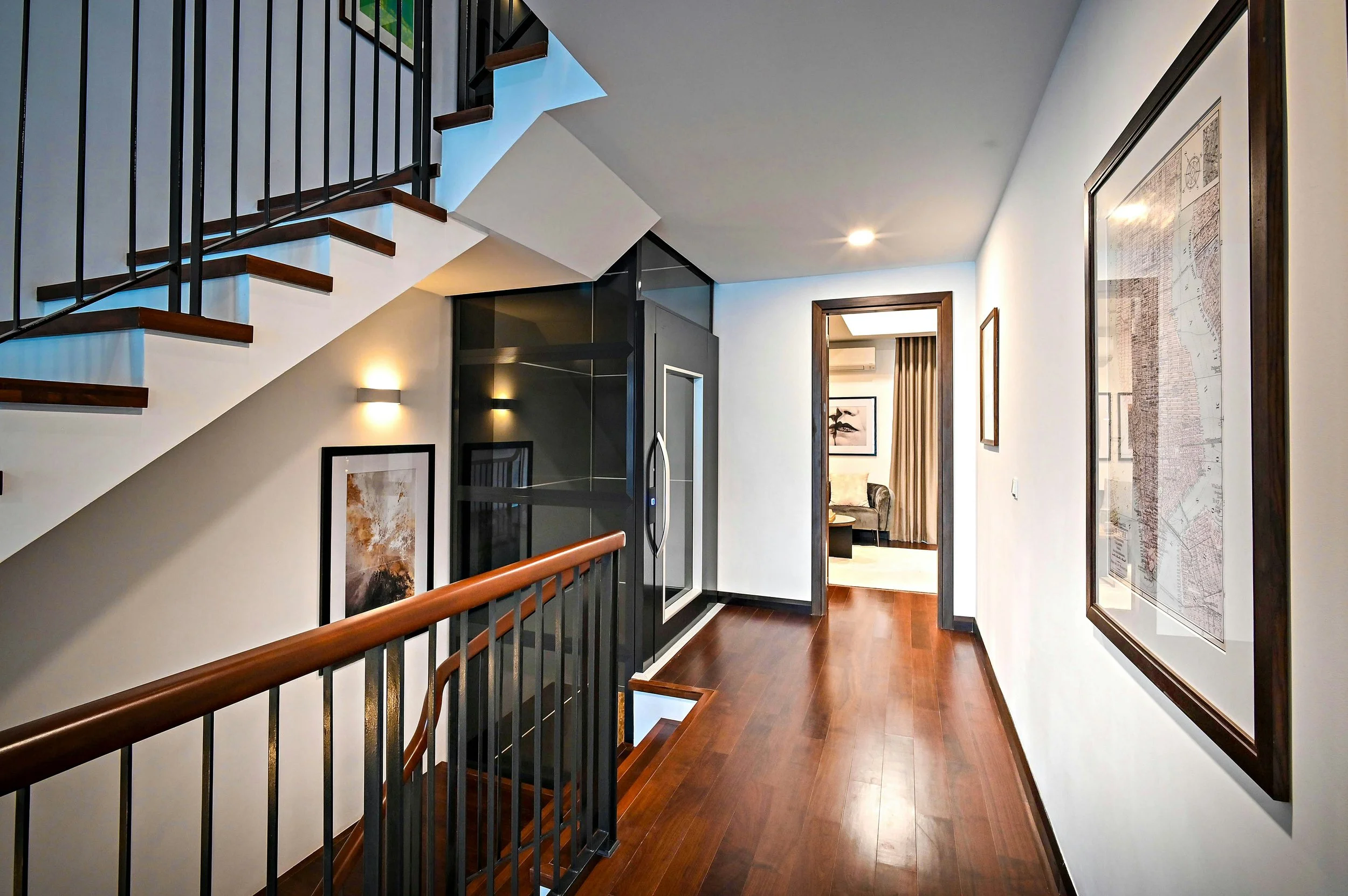 Orange County California remodel Interior view of a modern staircase and hallway in a home, featuring wooden flooring, black metal railings, framed artwork on white walls, and a doorway leading to a room with curtains.