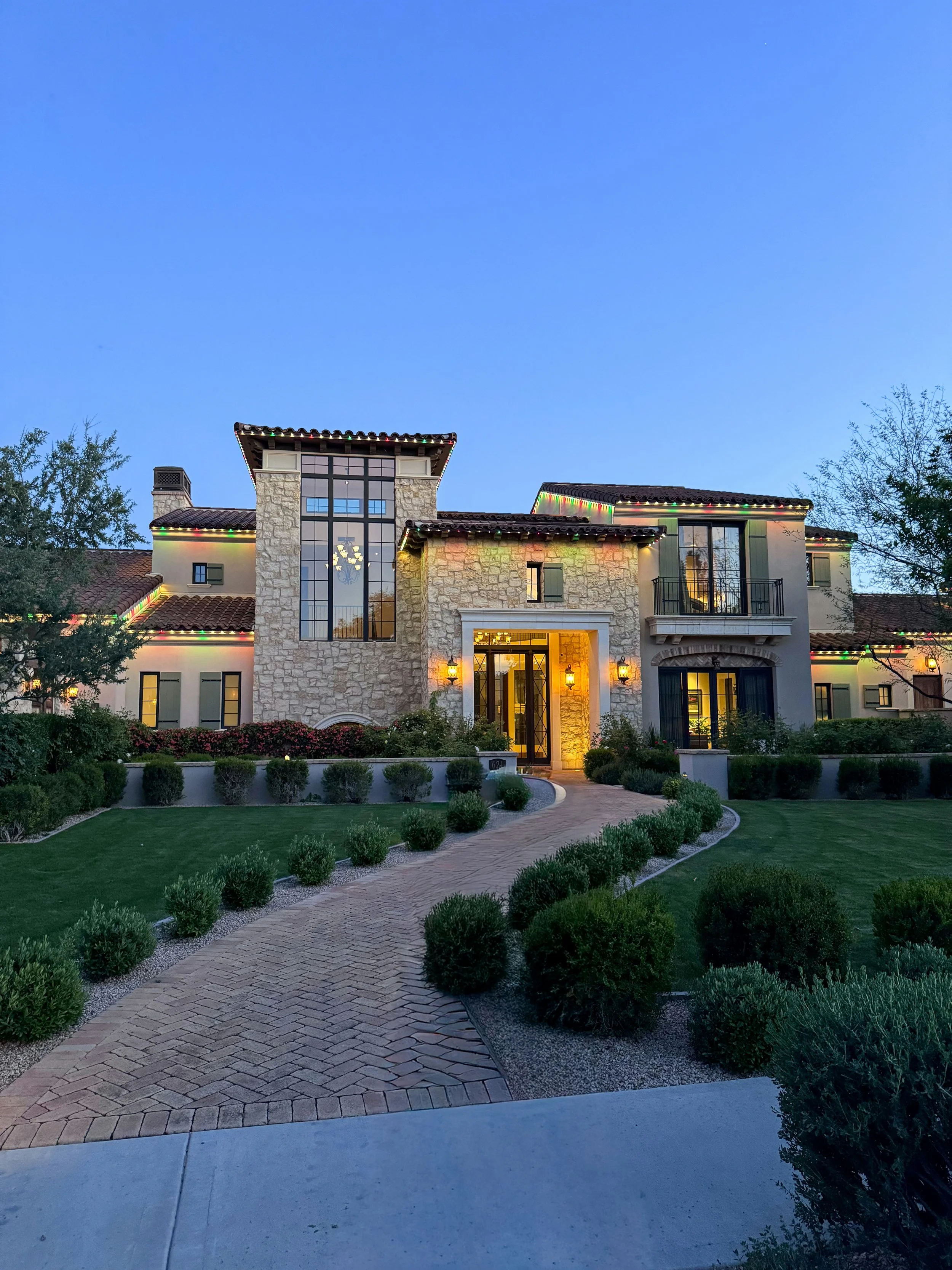 A large, custom, Southern California two-story house with stone and stucco exterior, string lights on roof edges, front yard with a curved brick walkway, manicured bushes, and trees, illuminated with warm lighting inside and outside