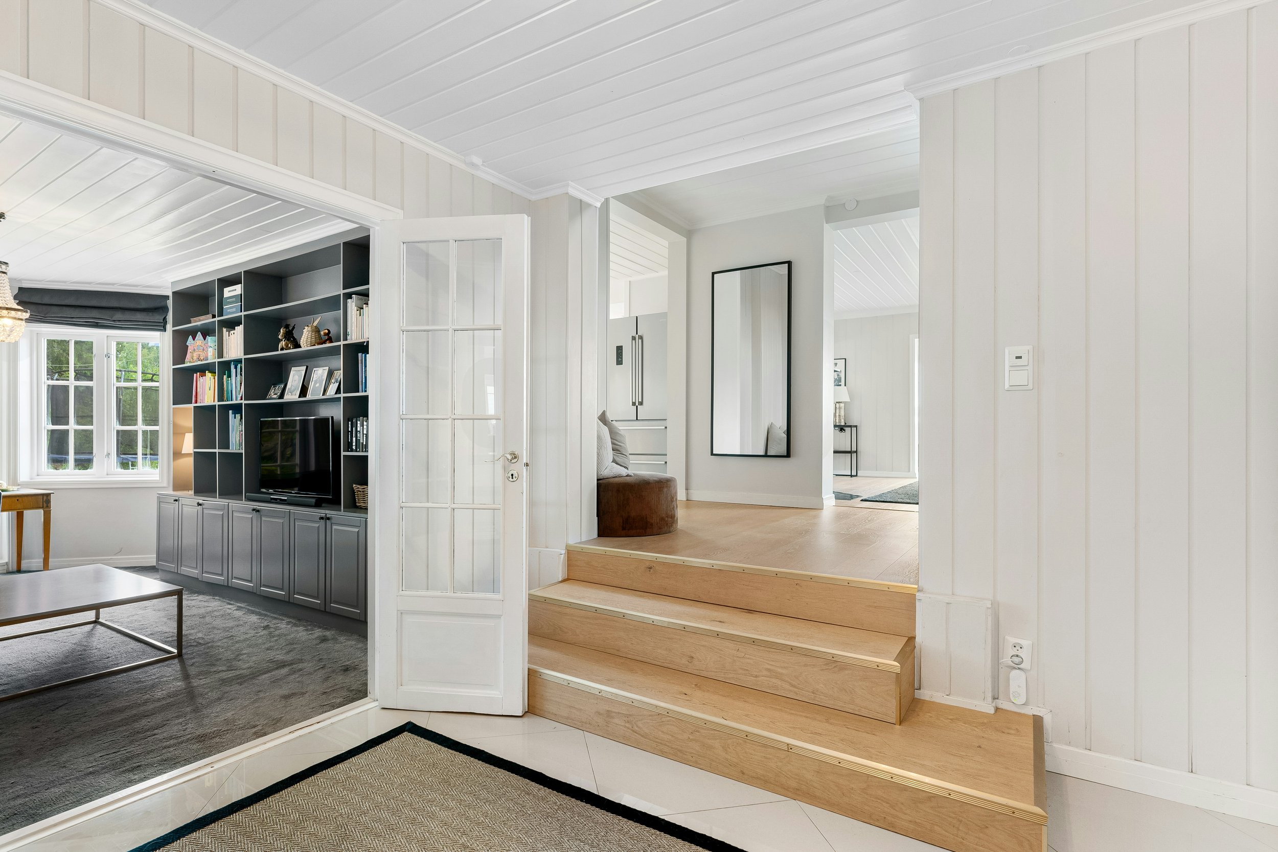 Orange County California remodel Interior view of a modern home with white paneled walls and ceiling, a staircase with wooden steps, and a living area with built-in dark gray shelving and a window with dark gray blinds.