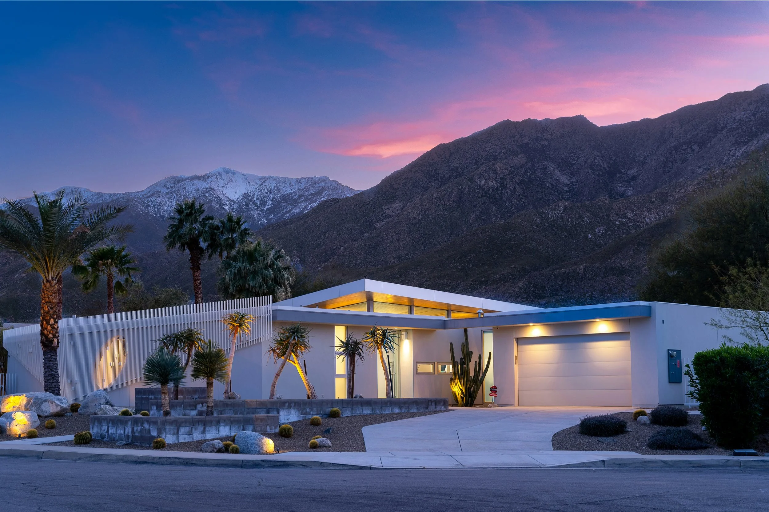 Southern California modern house with white exterior, large windows, and flat roof, surrounded by desert landscaping including palm trees, cacti, and decorative rocks, set against a mountain range at dusk with a colorful sky.