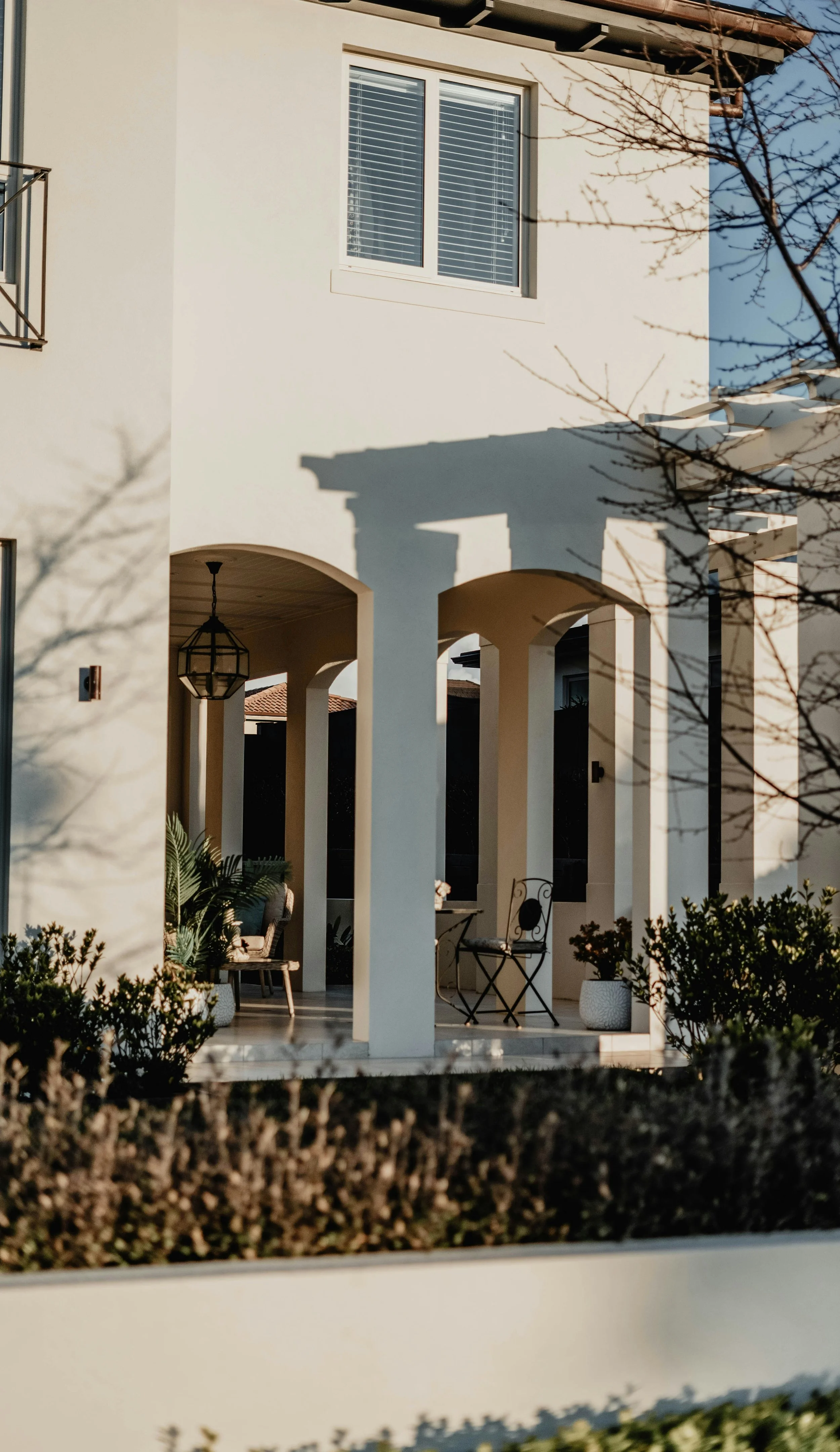 Orange County California custom home exterior, white house with a front porch featuring arched openings, outdoor seating, potted plants, and a hanging lantern. The house has a window with white blinds and shadows cast by nearby trees.