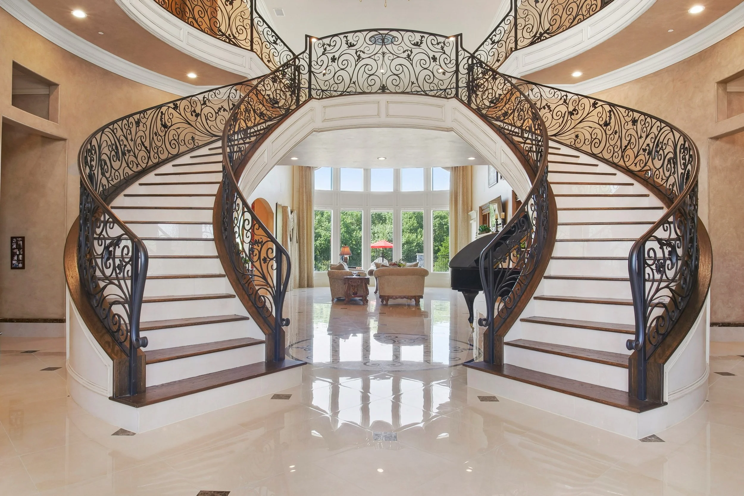 Orange County California remodel Interior of a grand home with a sweeping double staircase with ornate wrought iron railings, leading to a balcony overlooking a living area with large windows and outdoor scenery.