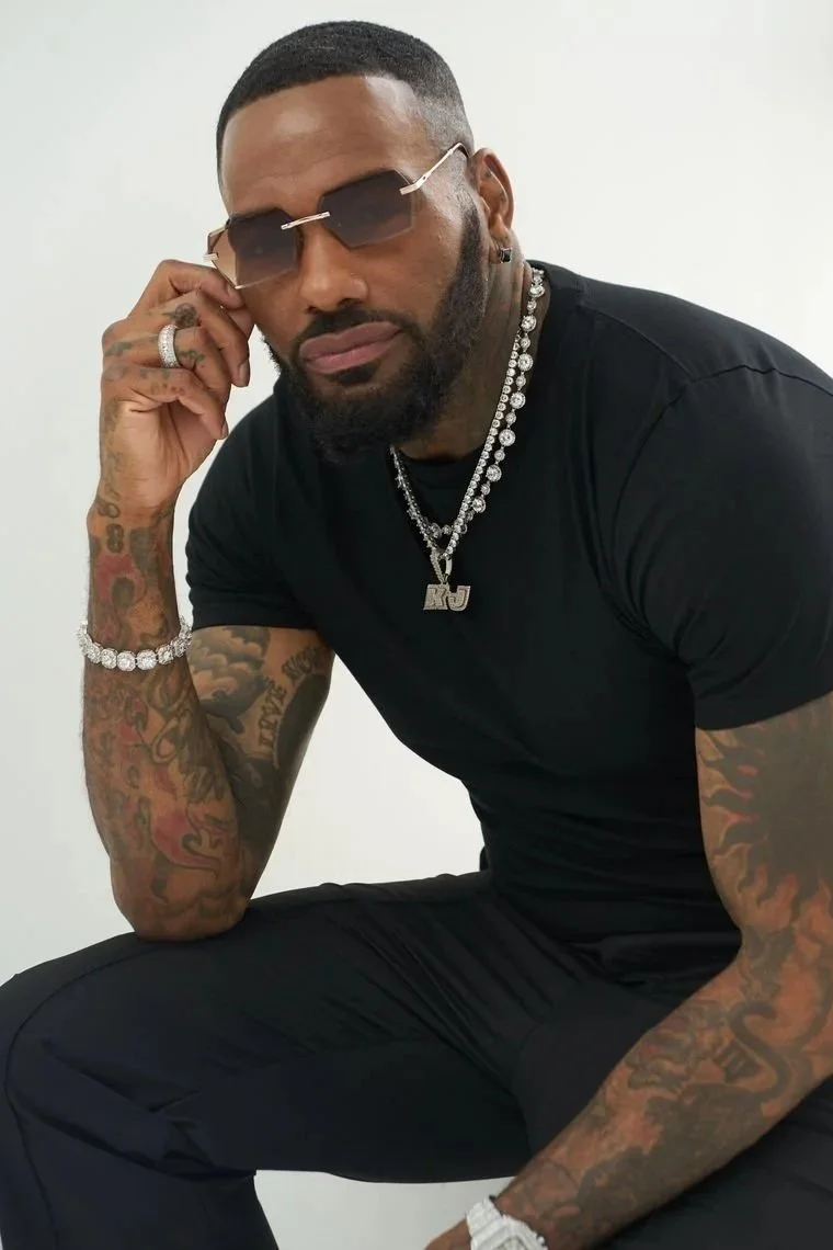 A man with short hair and a beard wearing dark sunglasses, multiple necklaces, a black t-shirt, and black pants, sitting against a plain white background.