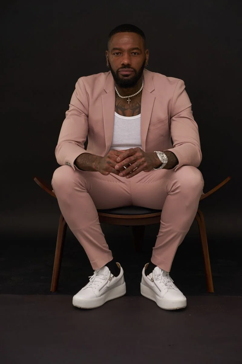 Man sitting on a wooden chair against a black background, wearing a pink suit, white sneakers, a white tank top, a pearl necklace, and a watch.