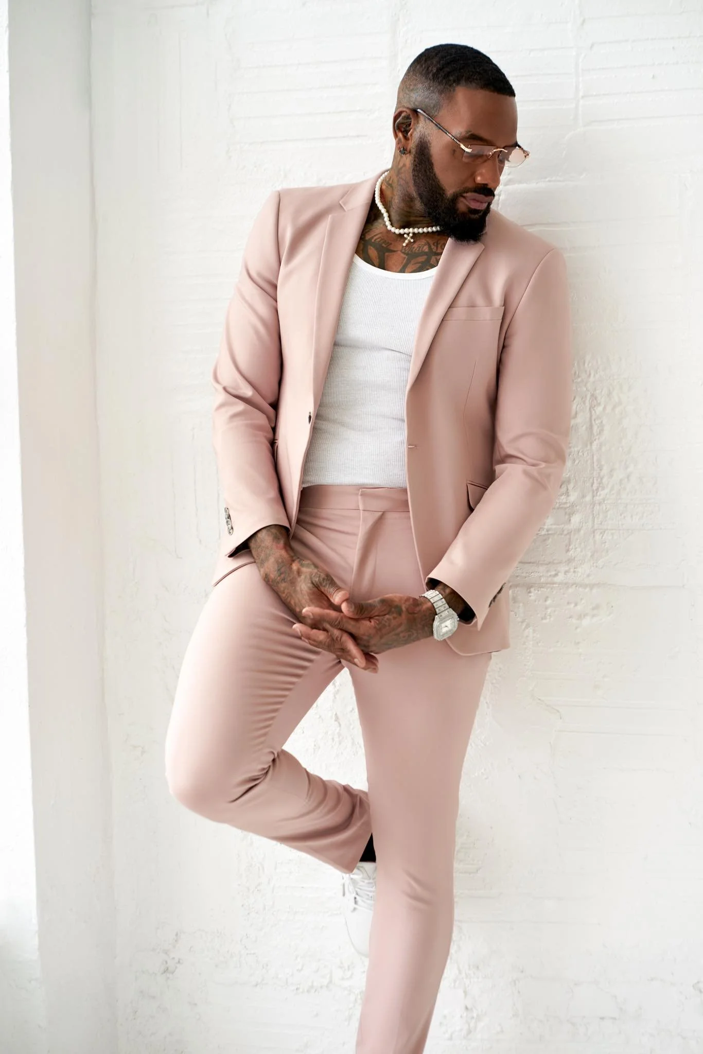 A man with glasses and a beard dressed in a light pink suit, white tank top, and white sneakers, standing against a white brick wall, with one hand covering his private parts and the other resting on his hip.