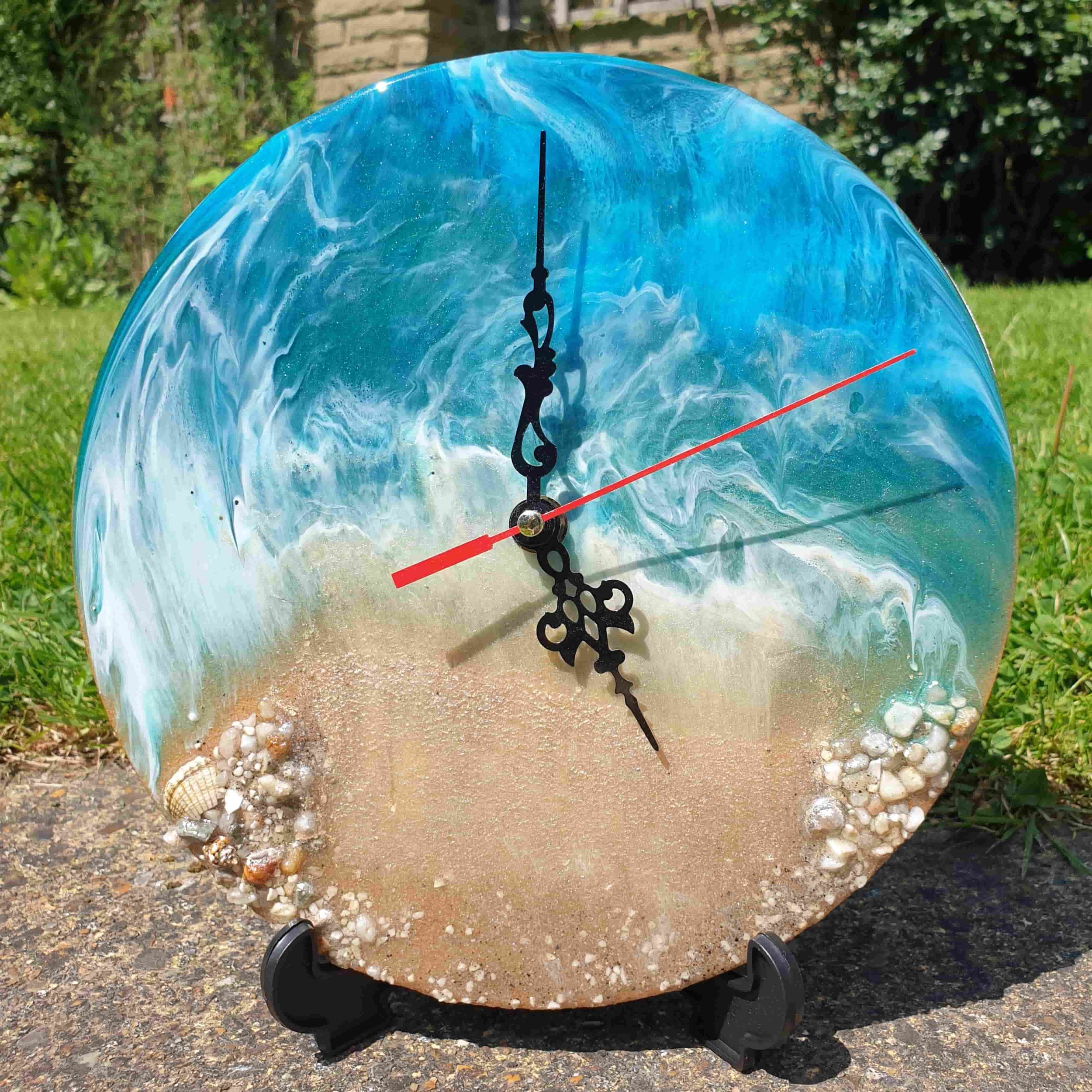 A decorative clock with a blue marbled face, featuring black hour and minute hands, and a red second hand, set outdoors on a stand with a background of green grass and bushes.