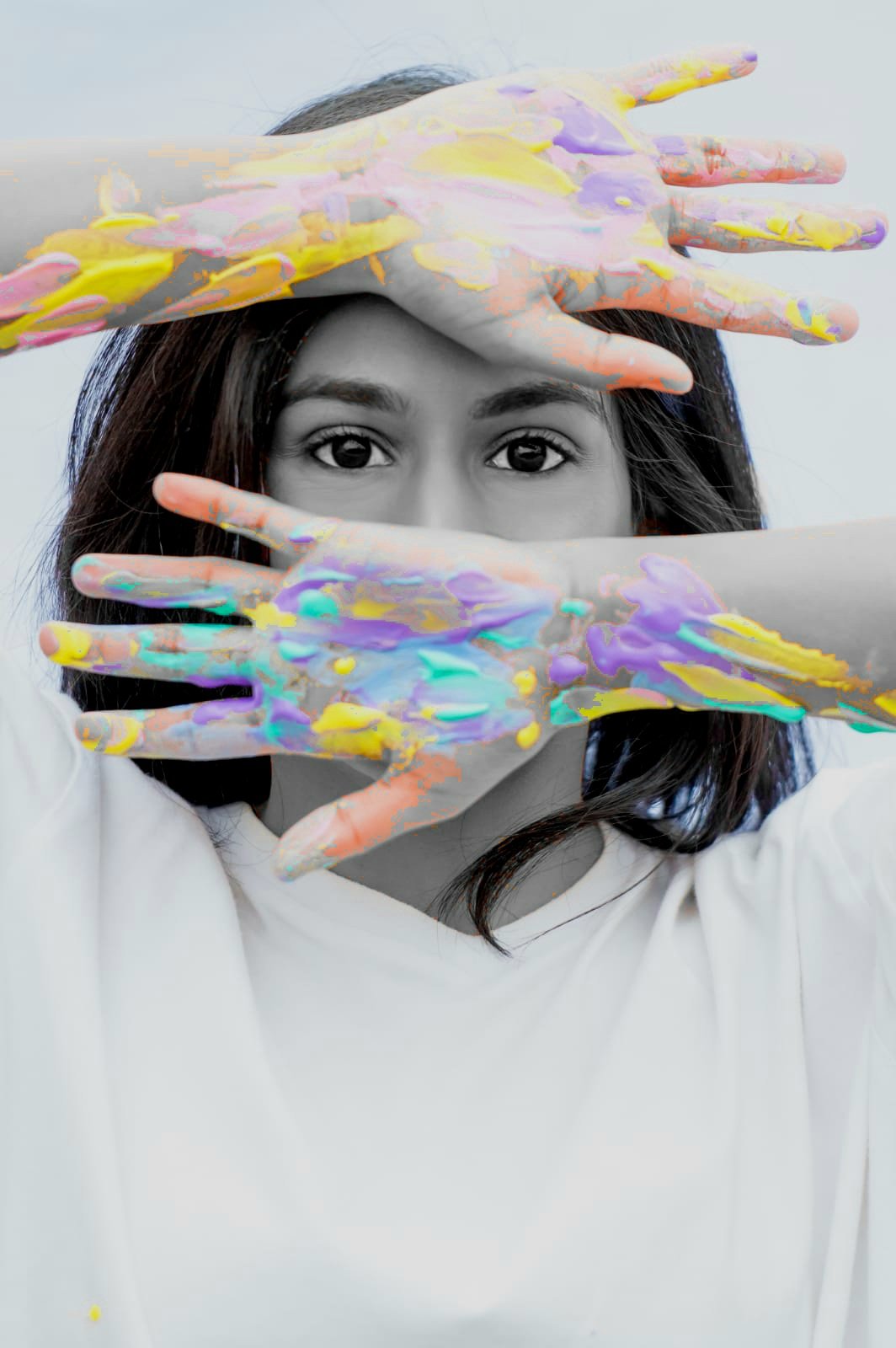 A woman with dark hair looks directly at the camera through her hands covered in colorful paint, creating a striking contrast with the black and white background and her monochrome clothing.