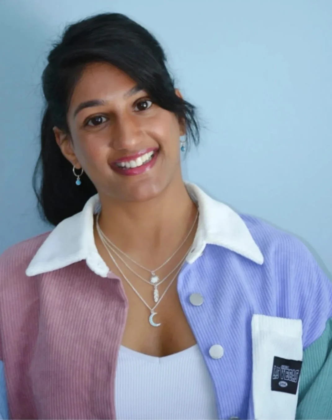 Smiling woman with dark hair, wearing layered necklaces, earrings, and colorful jacket over a white top, against a light blue background.