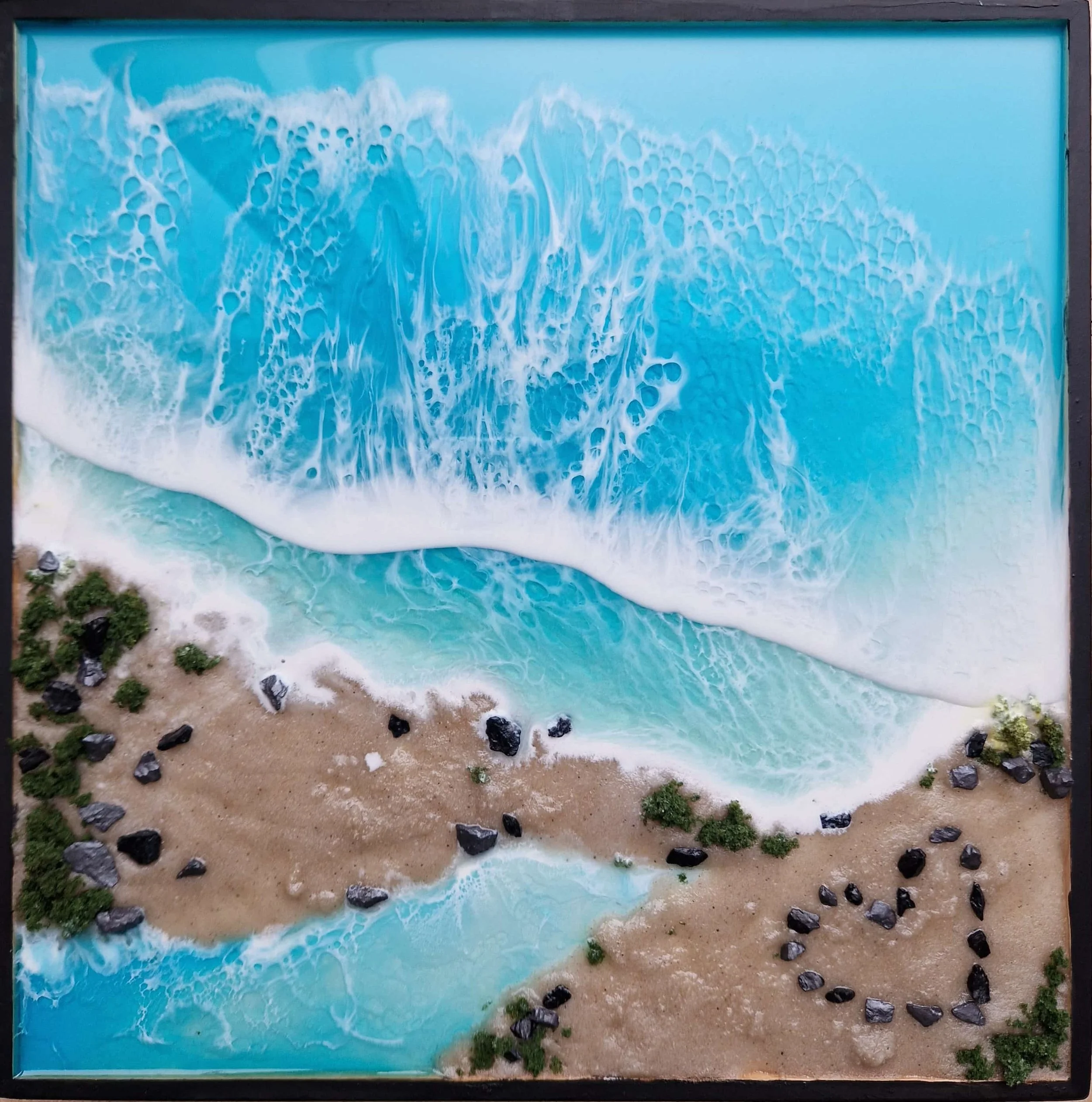 Model of a beach scene with turquoise water, white waves crashing onto a sandy shore, small rocks along the beach, and patches of greenery.