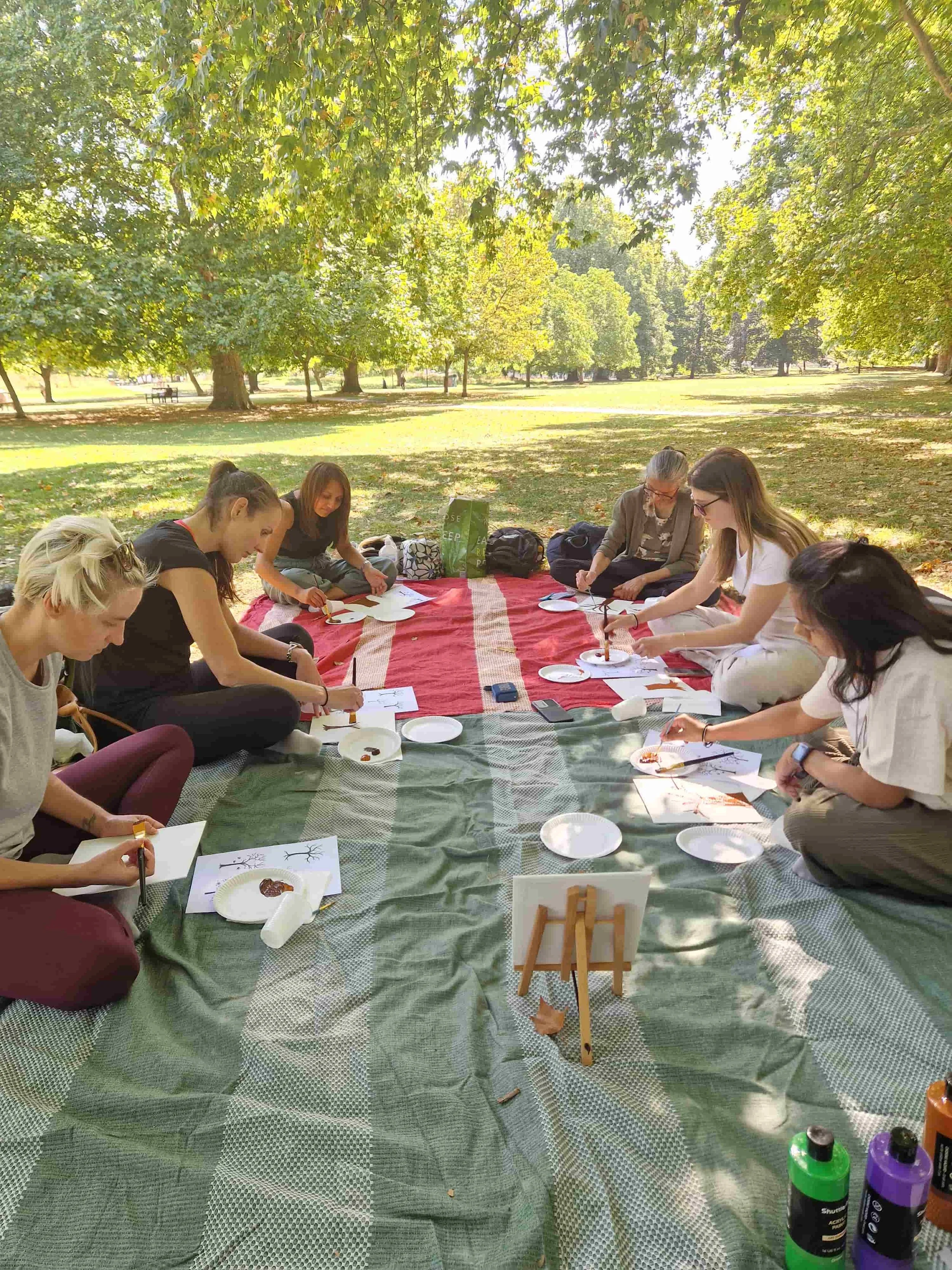 Group of people sitting in a circle on a picnic blanket in a park, engaged in a painting or craft activity under shade of trees.