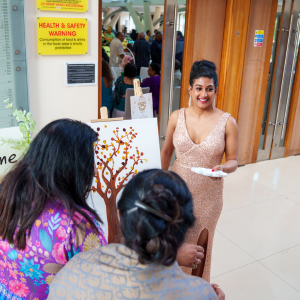 A woman in a sparkly dress holding a paper, smiling and talking to two women at an event or gathering inside a building. Other people are visible in the background.