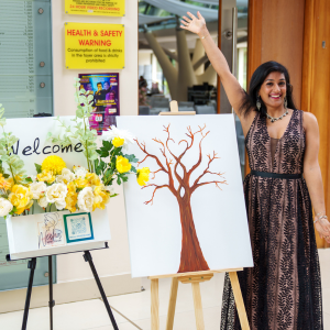 A woman in a black and brown dress smiling and waving next to a painted tree on an easel and a welcome sign with yellow flowers in a vase, at an indoor or outdoor event.