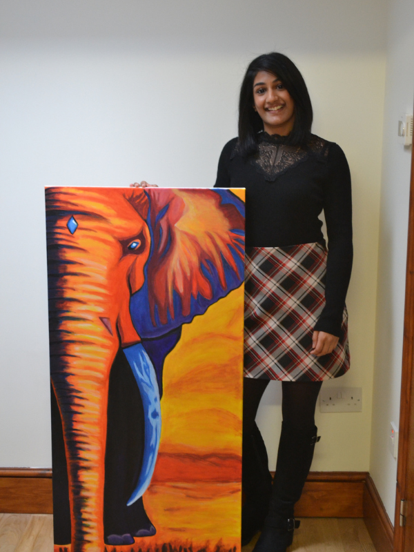 A woman with black hair, wearing a black top with lace details, a plaid skirt, and black leggings, standing indoors beside an orange and blue elephant painting.