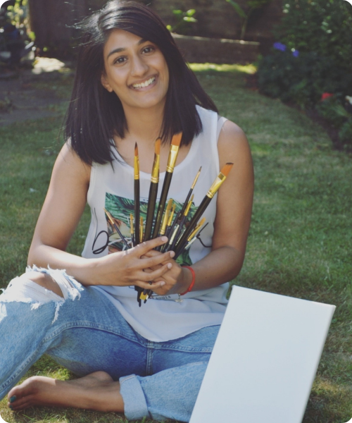Young woman sitting on grass outdoors with a laptop, holding paintbrushes, smiling at the camera.