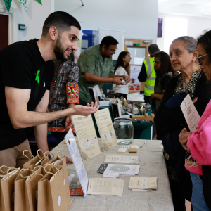 Man with a beard talking to women at a booth with informational materials and brown paper bags, at an indoor event with other people and vendor tables.