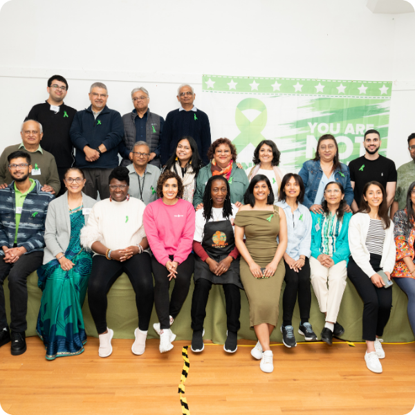 Group photo of diverse adults and seniors in a room with a green awareness ribbon on their shirts, standing and sitting in front of a green banner with a white awareness ribbon and text, indicating a gathering or event related to awareness or advocacy.