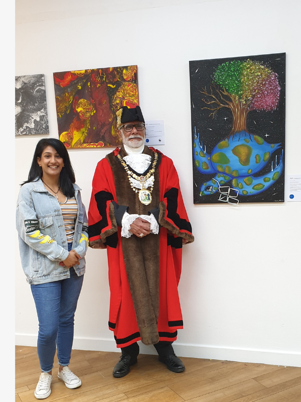 A young woman in a denim jacket and striped shirt standing next to a man dressed in a ceremonial robe with medals and a hat, at an art exhibition with colorful paintings on the wall behind them.