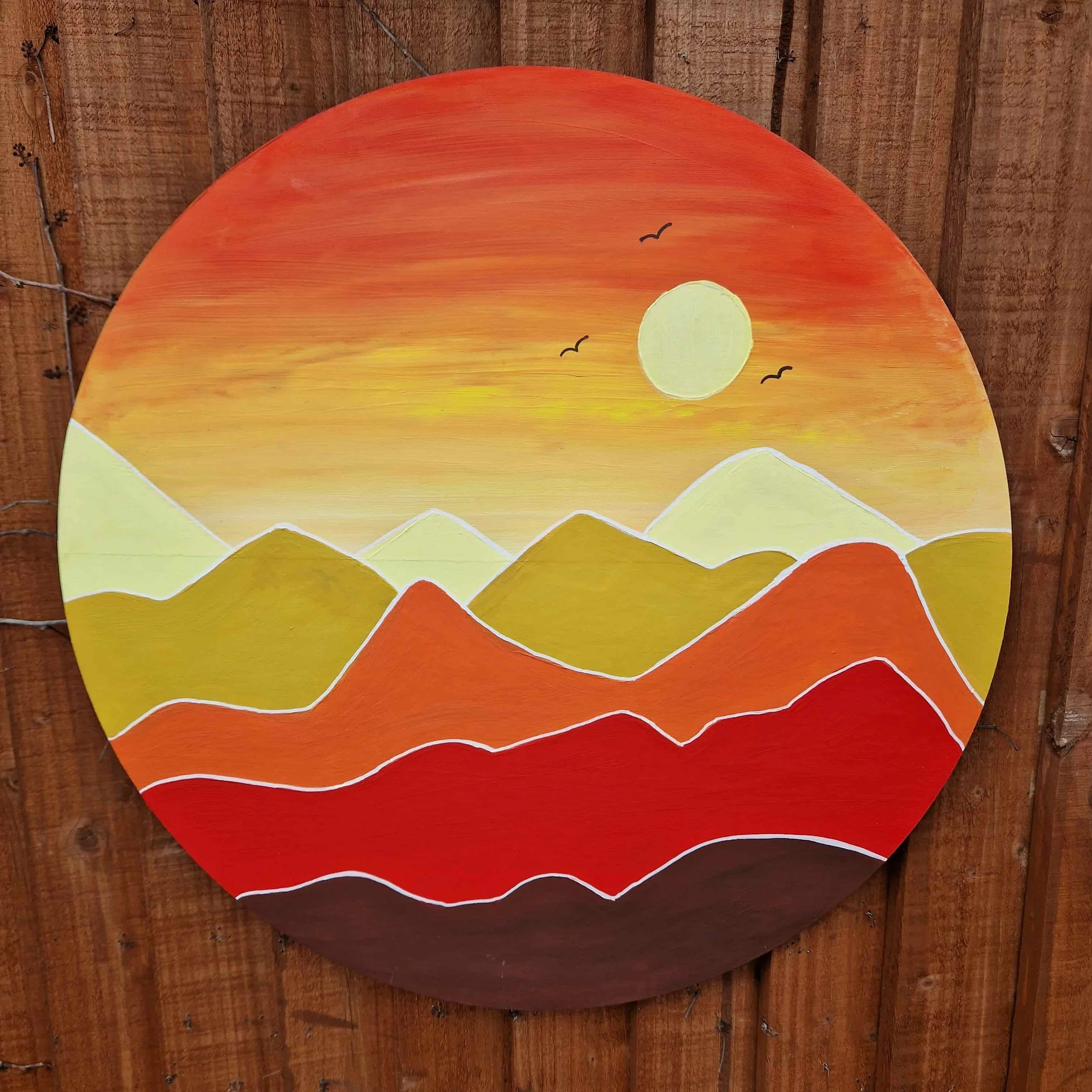 Colorful, semi-circular wooden fish-shaped sign with a sunset design, including a yellow sun and black birds, mounted on a wooden wall.