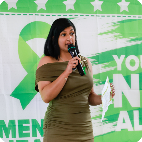 Woman speaking into a microphone at an event with a green sparkle-themed backdrop reading "YOUNG WOMEN IN ALL".