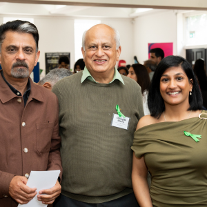 Three diverse adults at a social event, smiling, wearing green ribbon pins, indoors with other attendees in the background.