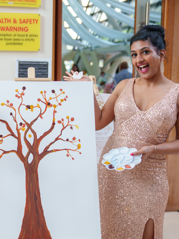 Woman in a sparkling gold dress standing next to a painting of a tree, holding a paint palette with various colors and smiling.