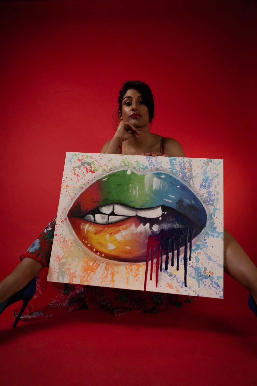 A woman with dark hair and red lipstick sitting in front of a red background, holding a colorful painting of lips with black dripping paint.