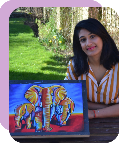 A woman with black hair wearing a striped yellow and white shirt sitting outdoors at a wooden table with a colorful elephant painting in front of her, in a green garden with trees and flowers in the background.