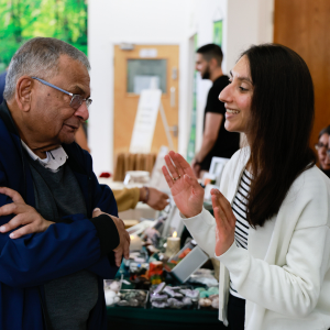 A young woman and an elderly man are smiling and talking at an indoor event or market, with other people and tables in the background.