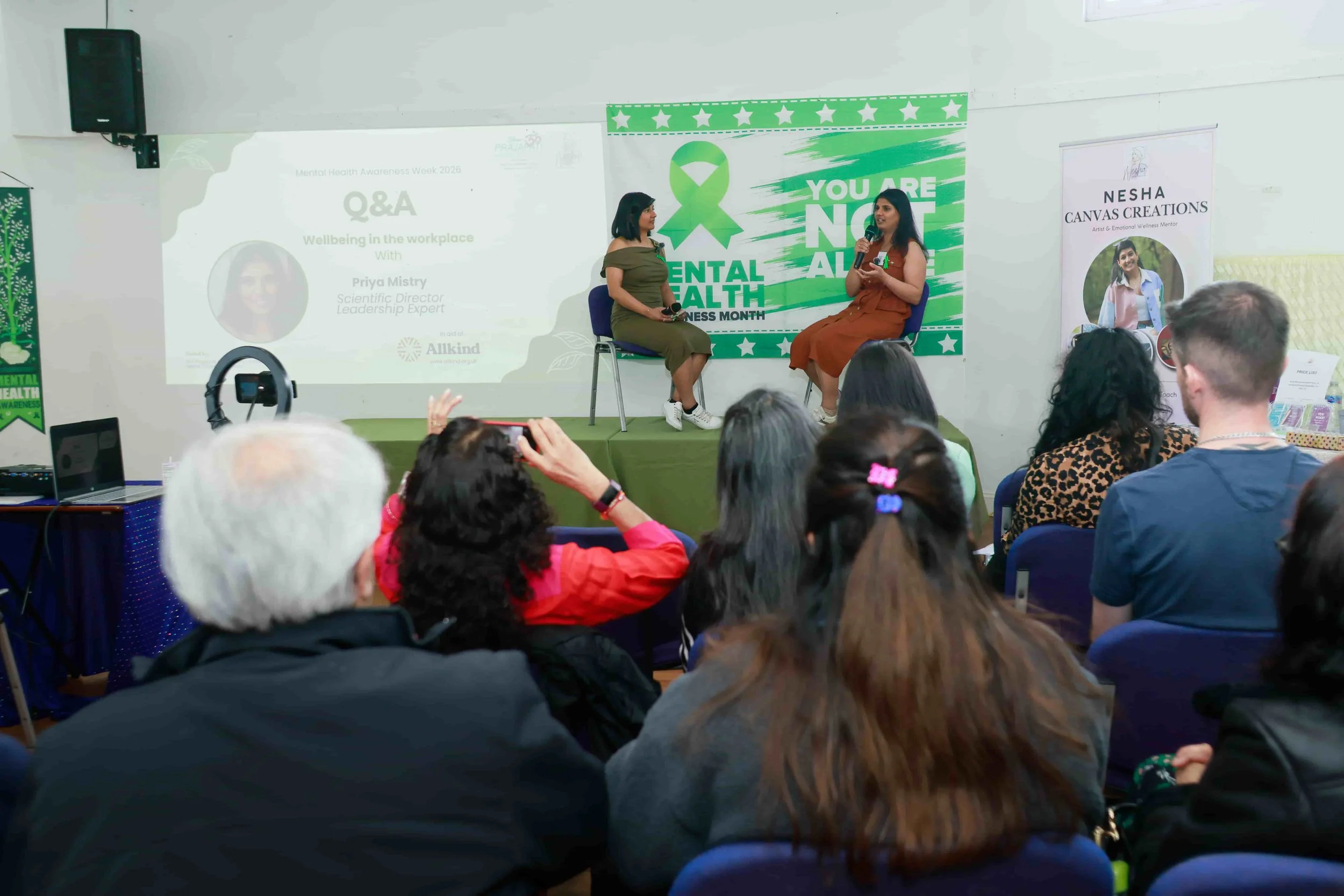A woman in a green dress and a woman in an orange dress sitting on stage during a mental health awareness event. Audience members are watching and taking photos. A large banner behind them displays a green ribbon and the phrase "YOU ARE NOT ALONE" related to mental health awareness.