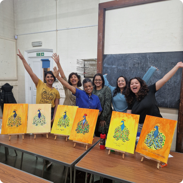Group of five women in a classroom with paintings of peacocks on easels, celebrating their art projects.
