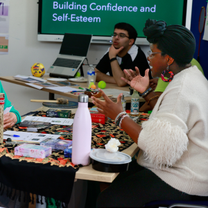 Three people sitting around a table during a discussion, with a colorful background that includes a screen displaying 'Building Confidence and Self-Esteem'.