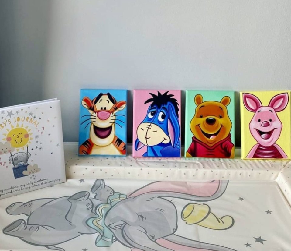 Four colorful blocks featuring cartoon characters from Winnie the Pooh, including Tigger, Eeyore, Winnie the Pooh, and Piglet, are lined up on a shelf. To the left, there is a children's journal with a smiling sun and clouds on the cover, and a blanket or cloth with a sleeping animal, possibly a rabbit, and stars.