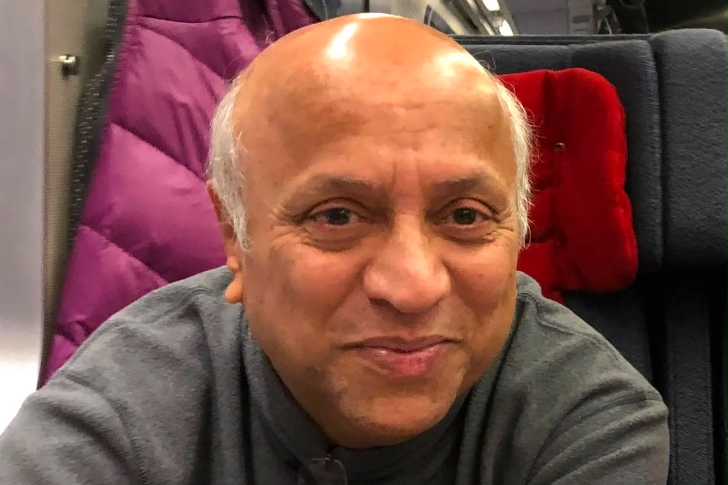 Neil's father smiling gently at the camera, seated on a train in a gray sweater with his glasses hooked on his collar.