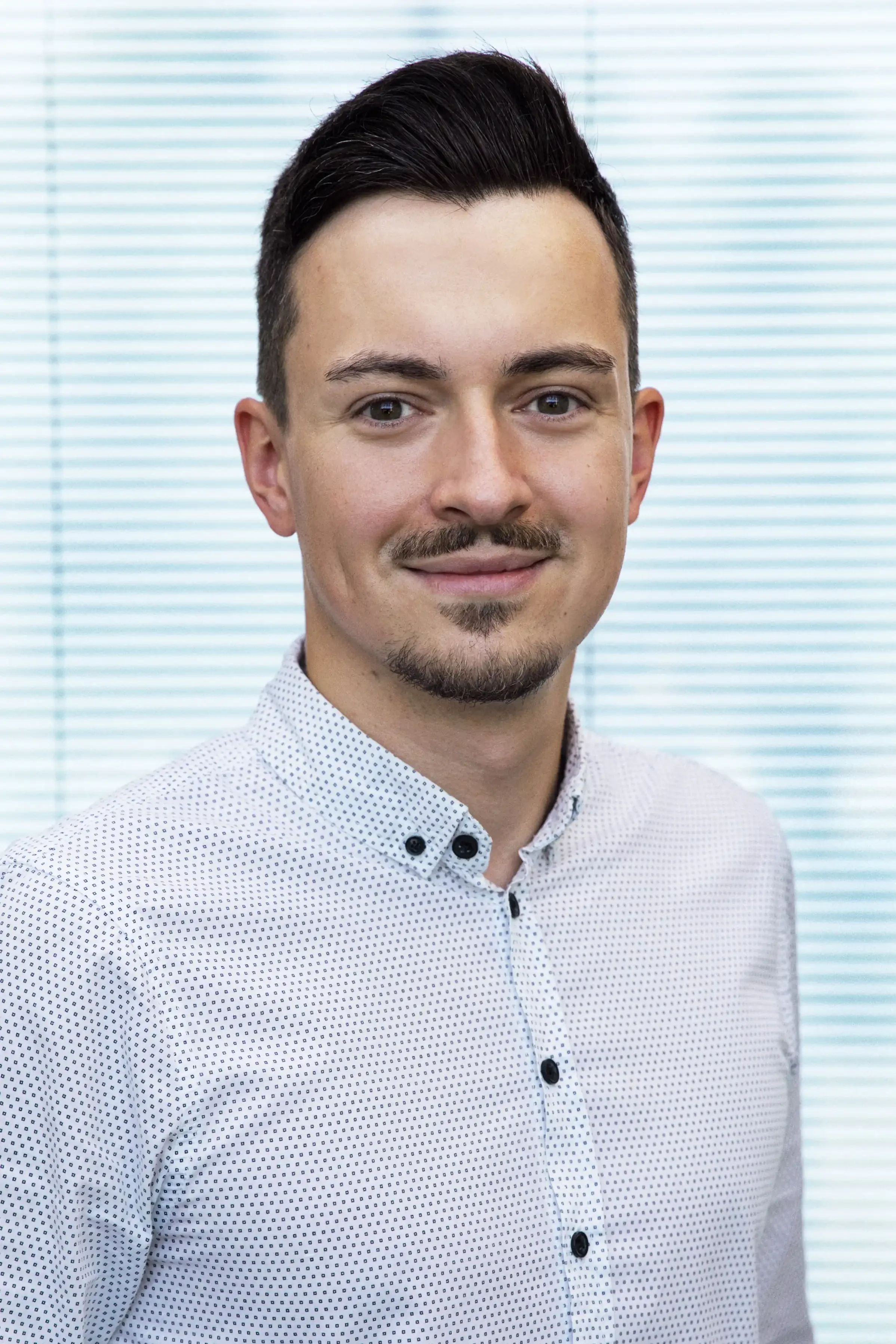 Ein Mann mit kurzen dunklen Haaren, leichter Gesichtsbehaarung, tragt ein hellblau-weiß gepunktetes Hemd, steht vor einem Hintergrund mit horizontalen blauen Fensterjalousien.