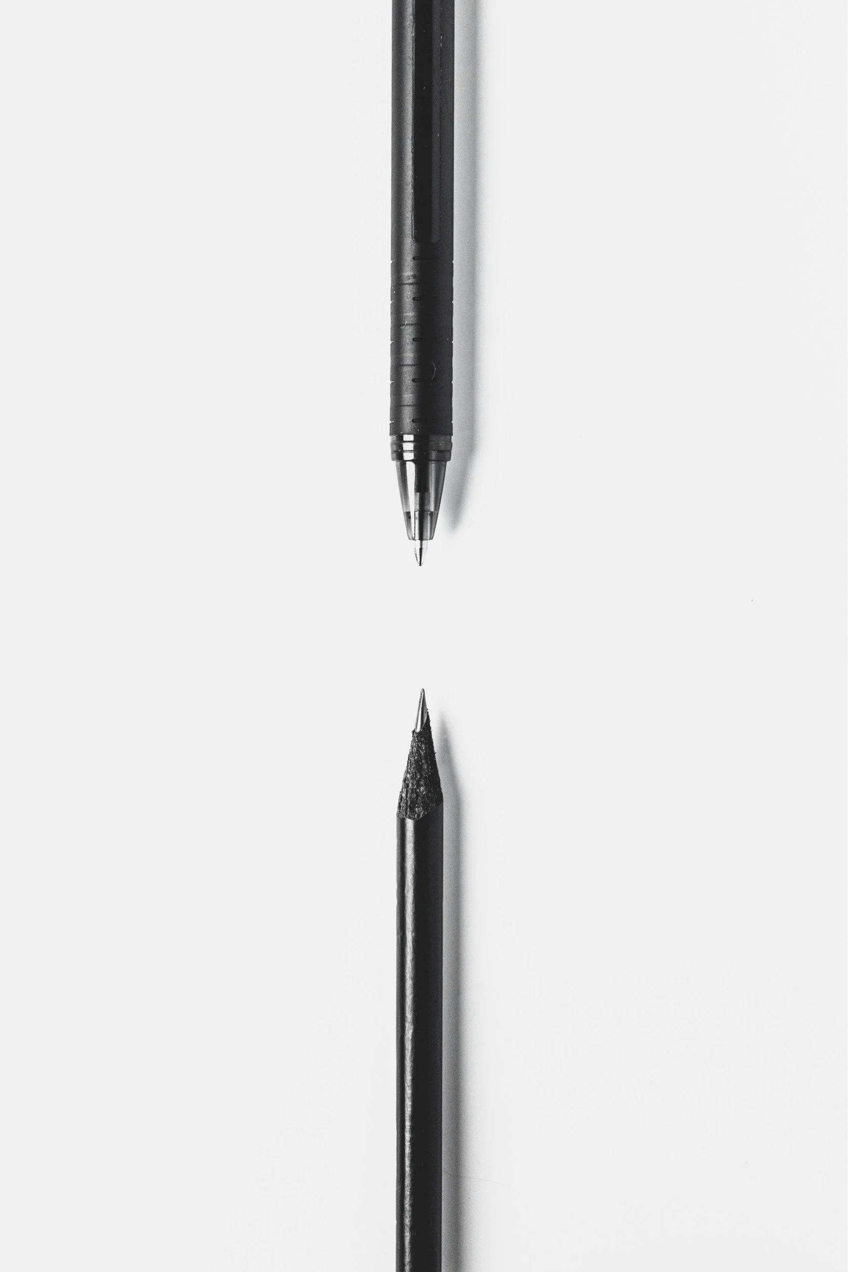 Close-up of two writing instruments, a black pen and a black pencil, facing downward on a white surface with their reflections visible.