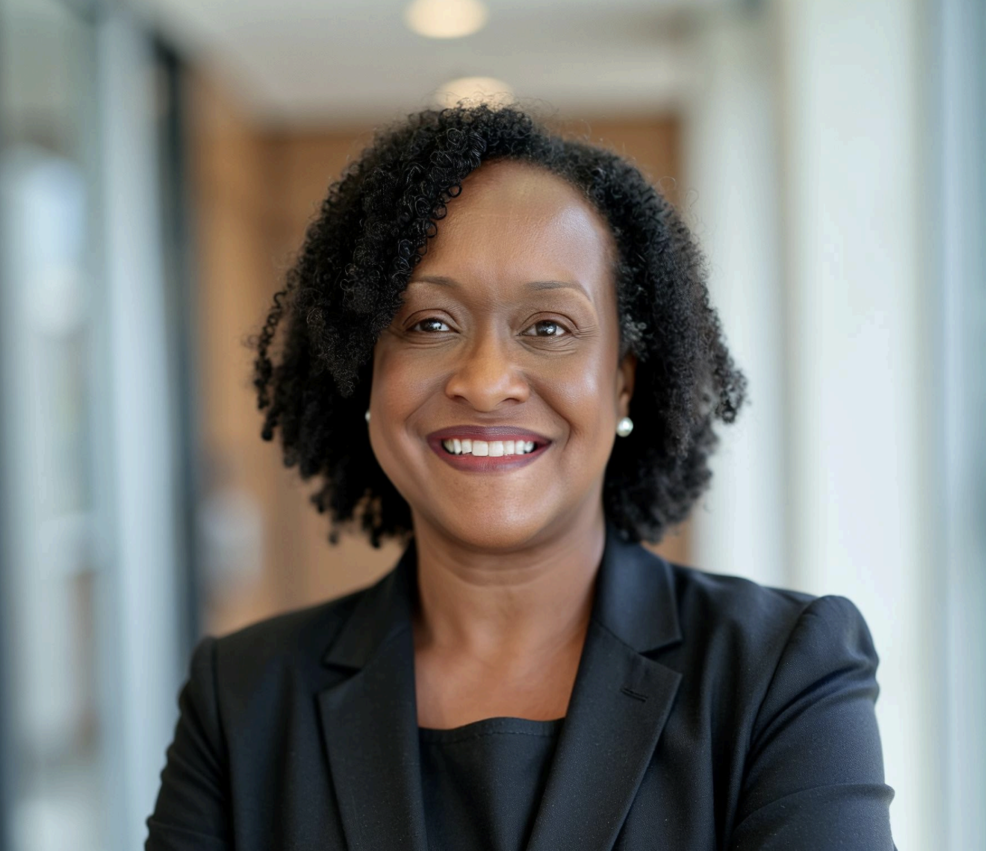 A smiling African-American woman with curly black hair, wearing pearl earrings and a black blazer, standing in a well-lit indoor environment.