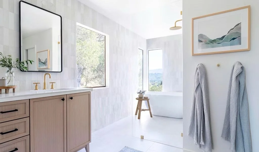 A modern bathroom with a large mirror, beige vanity with black handles, gold faucet, two gray towels hanging on the wall, a framed abstract art piece, a window with a tree outside, a bathtub with a small wooden stool, and a pitcher with flowers on the stool.