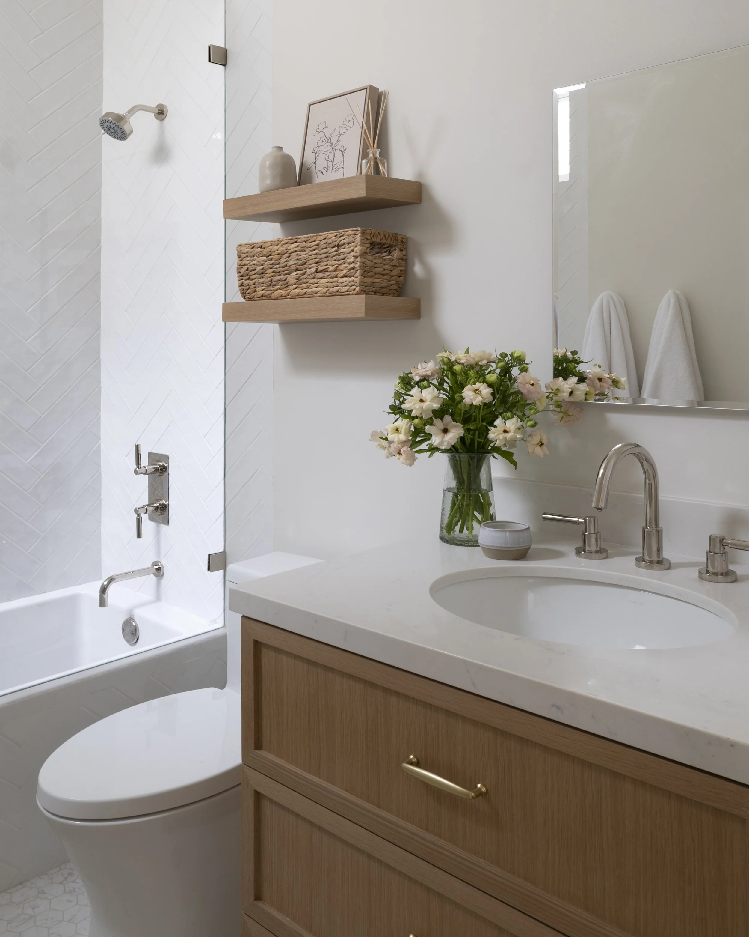 A modern bathroom with a white bathtub, a vanity with a wooden cabinet and a marble countertop, a vase of flowers, a large mirror, and wall shelves with decorative items. Two towels hang on the wall.