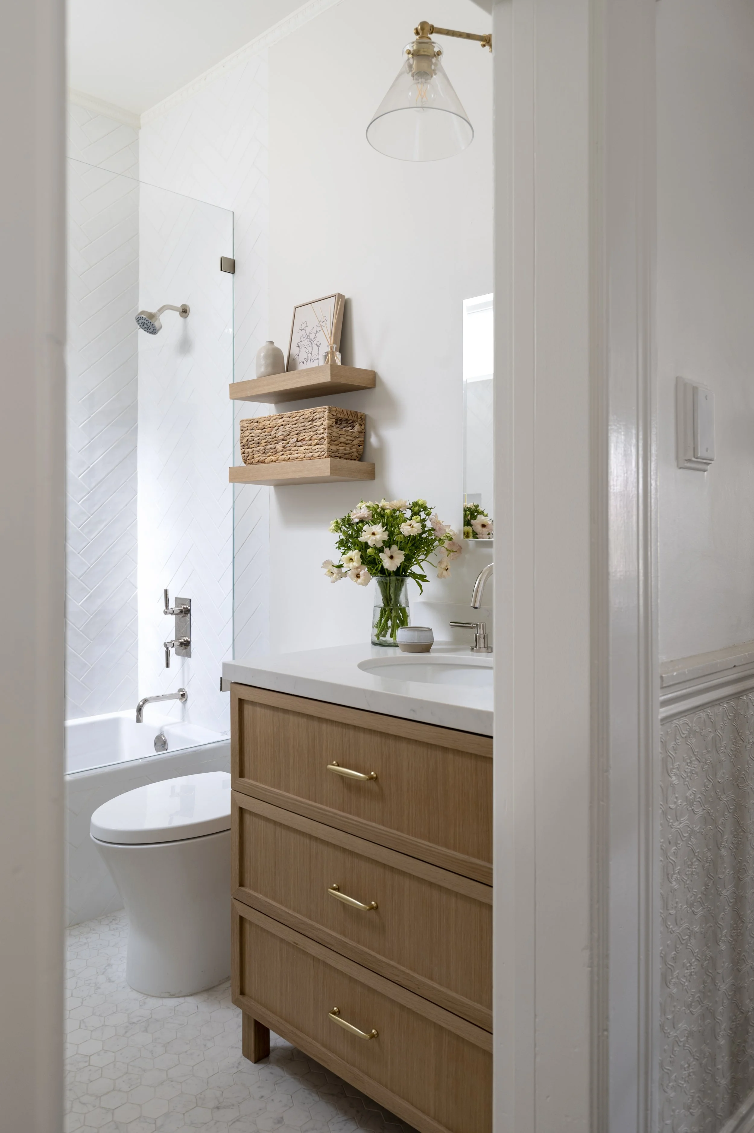 A modern bathroom with a white toilet, a wooden vanity with drawers, a vase of flowers on the vanity, floating shelves with decorative items, and a shower area with white subway tiles and a glass door.