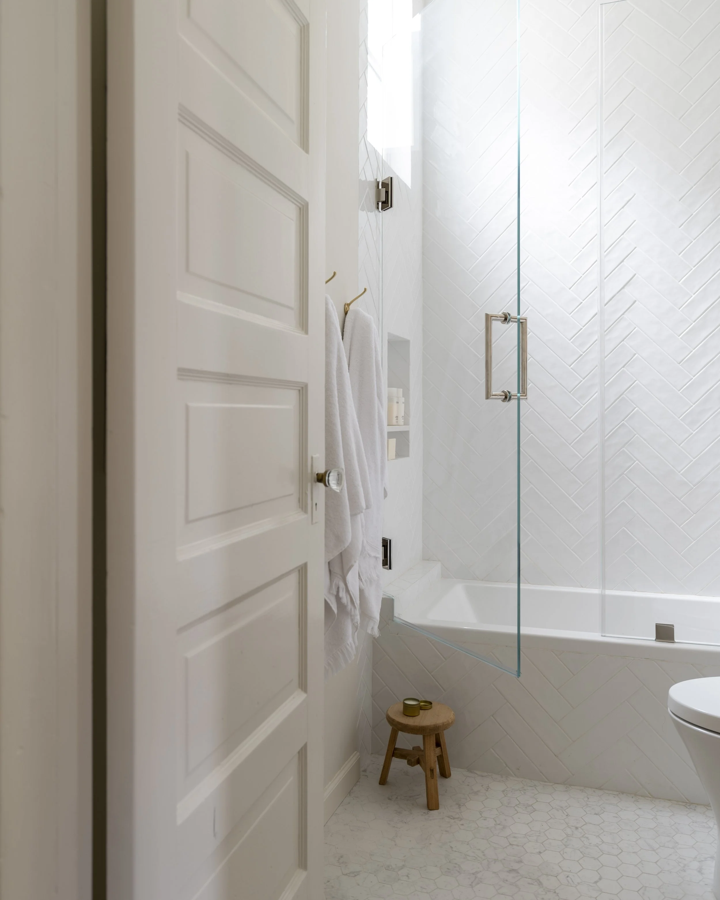 A bathroom with a white door, wall-mounted towel rack with white towels, a small wooden stool with a gold container, and a glass shower enclosure with a handle. The walls have textured white tiles, and the floor has hexagonal white tiles. Natural lig