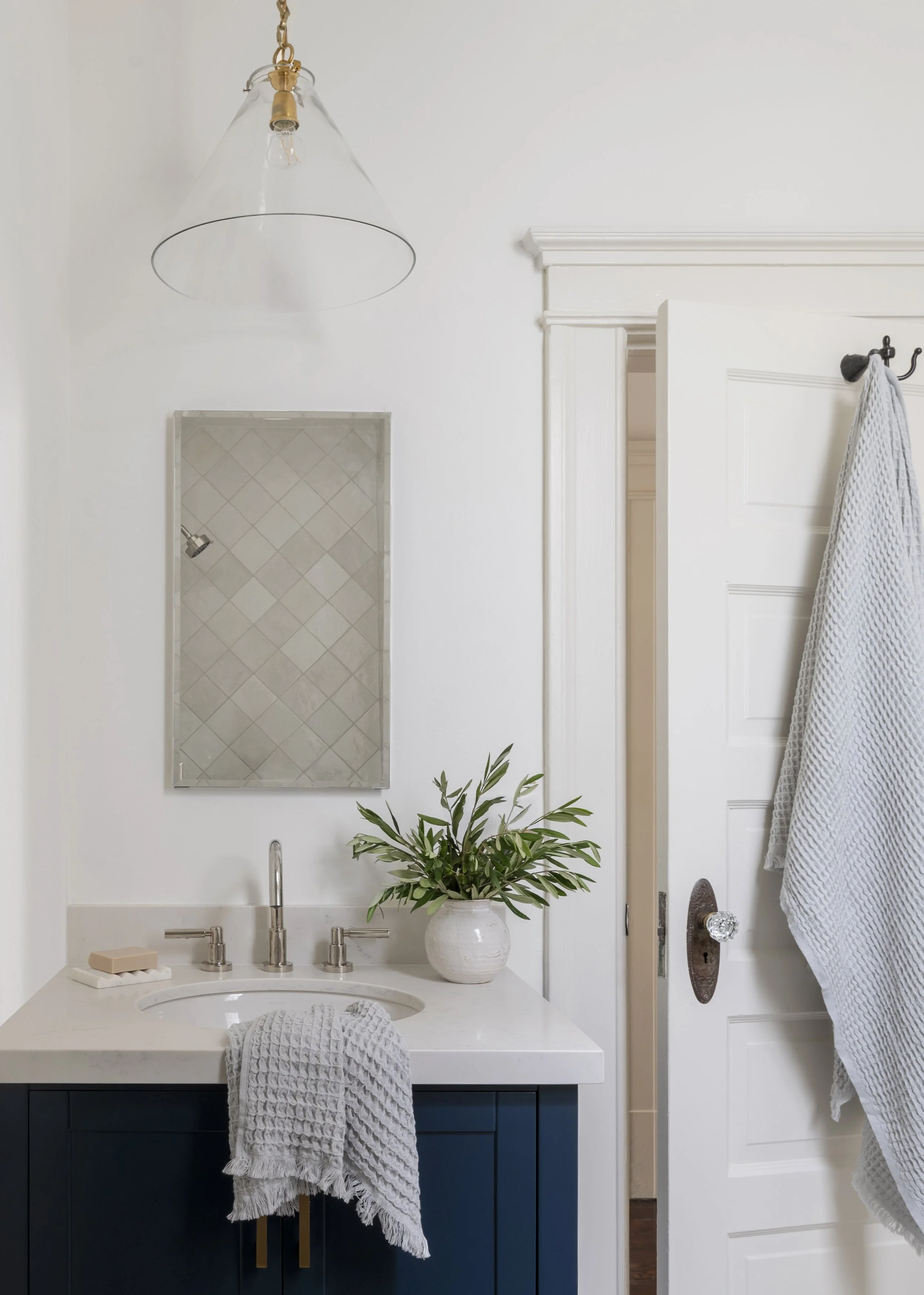 Bathroom vanity with a white countertop, a navy blue cabinet, a mirror, and a plant in a white vase. A soap bar, a towel, and a beige soap dish are on the countertop. A shower door and a white towel hanging on a hook are also visible.