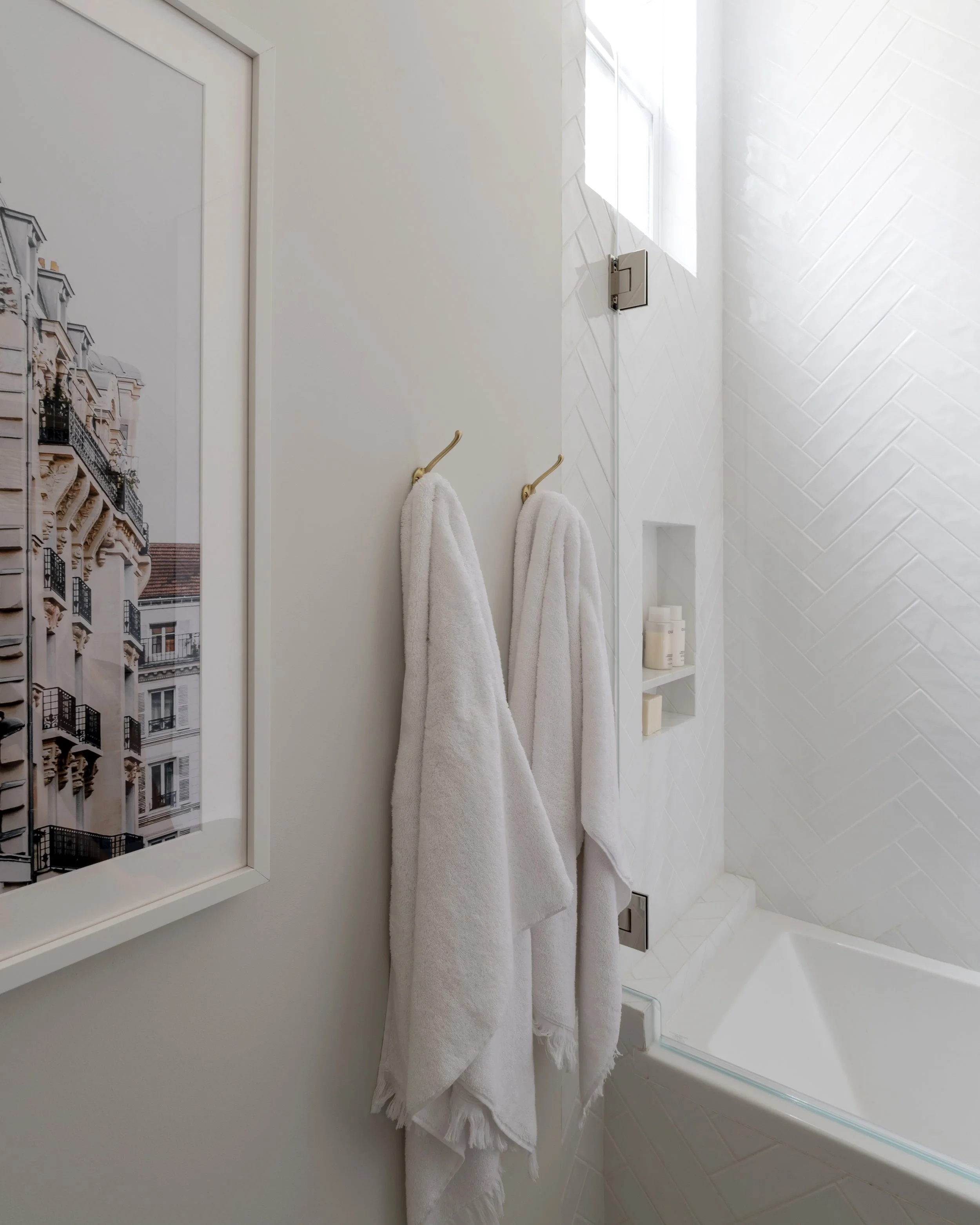 Interior view of a bathroom with two white towels hanging on gold hooks on a beige wall, a window with sunlight coming through, a partially visible bathtub, and a shelf with bottles, with a framed picture of a building on the wall.